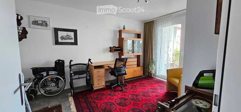 A furnished room with a desk, chair, shelf, sofa, wheelchair, mirror, picture frame, and plant. The room features a red patterned rug and large windows with curtains.