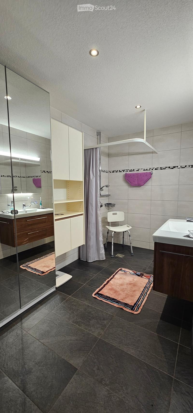 Modern bathroom with dark tiled floor, white walls, two sinks, a shower area with a curtain, a white chair, and a towel on the wall.