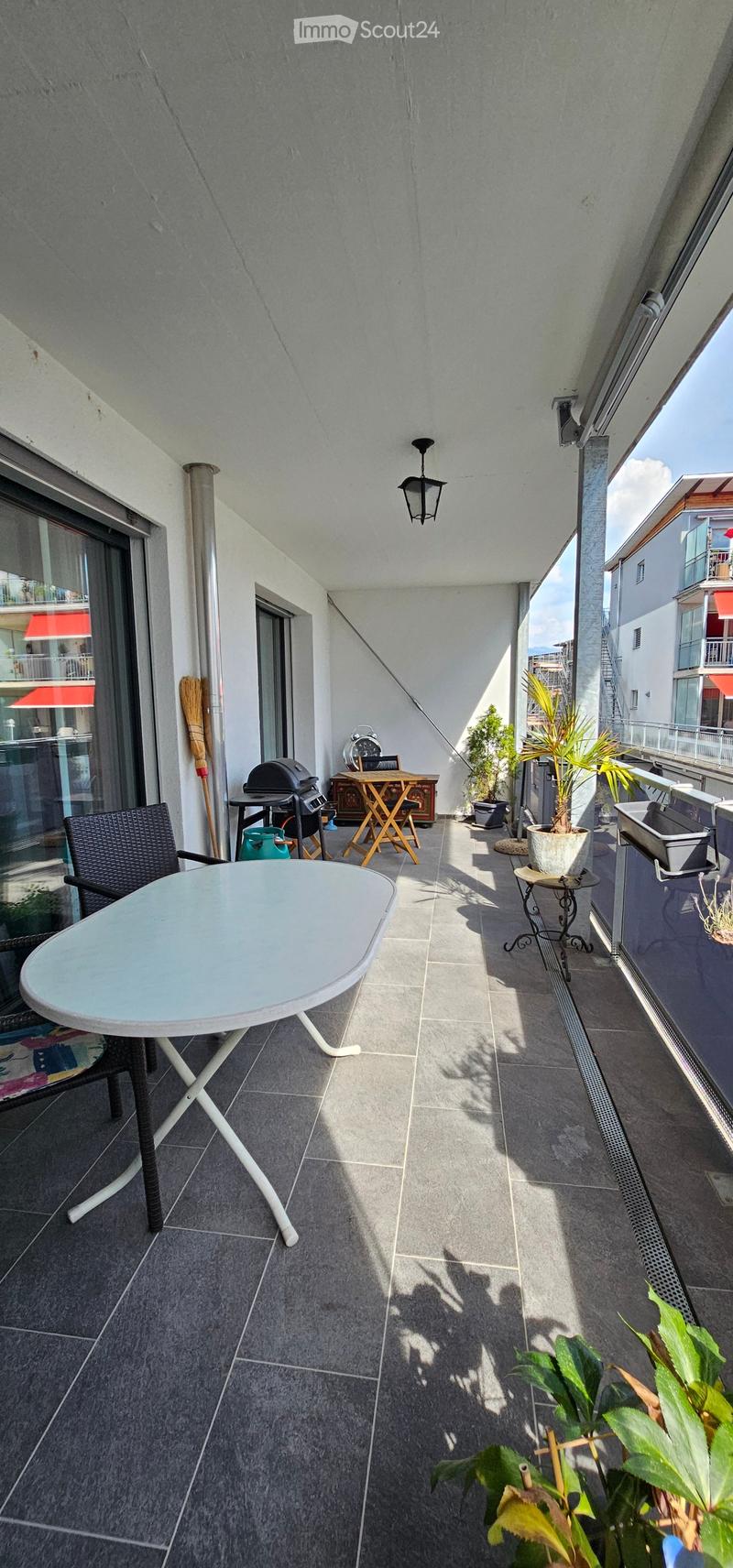 Balcony with tiled floor, white round table, black wicker chair, barbecue, small wooden table, potted plants, and a broom leaning against the wall.