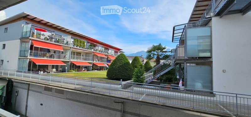 Modern multi-story building with balconies, red awnings, and a landscaped garden area.