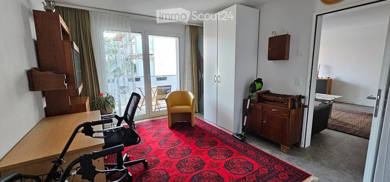 A furnished room with a red patterned rug, a yellow armchair, a black office chair, a wooden table, a white cabinet, and a small balcony area with outdoor furniture visible through the glass door.