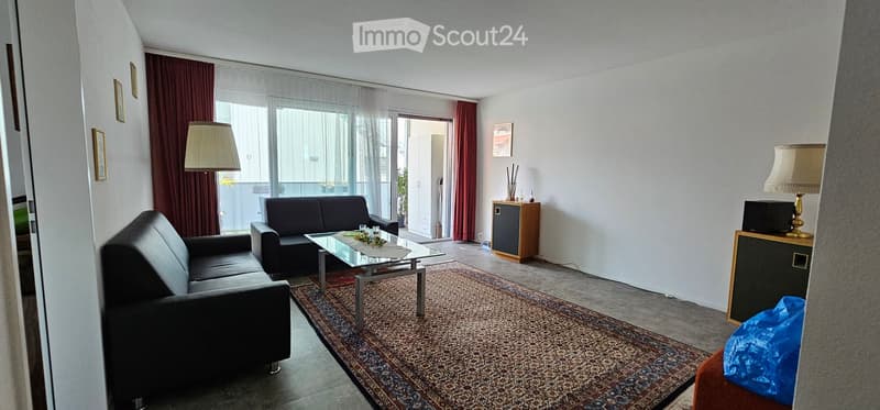 Modern living room with large windows and balcony access, featuring a grey sofa, glass coffee table, floor lamp, side table with TV, and decorative items.