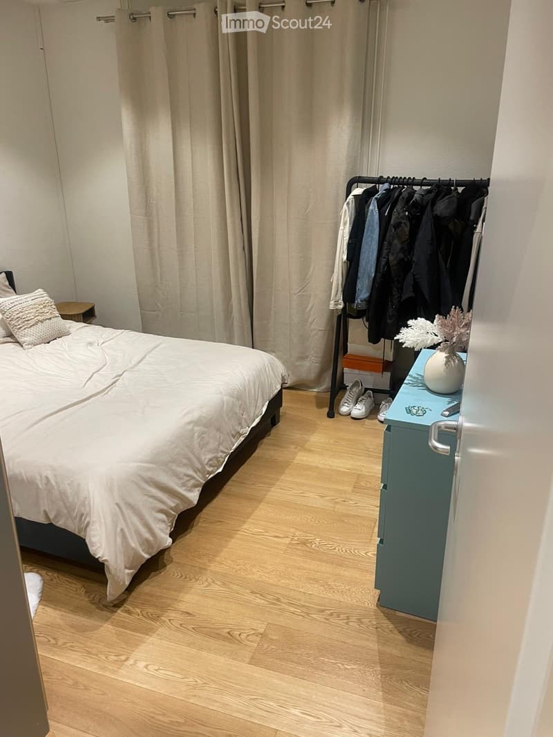 Cozy bedroom with a neatly made bed, a small nightstand, a clothing rack with hanging clothes, and a vase with decorative branches on a blue side table.