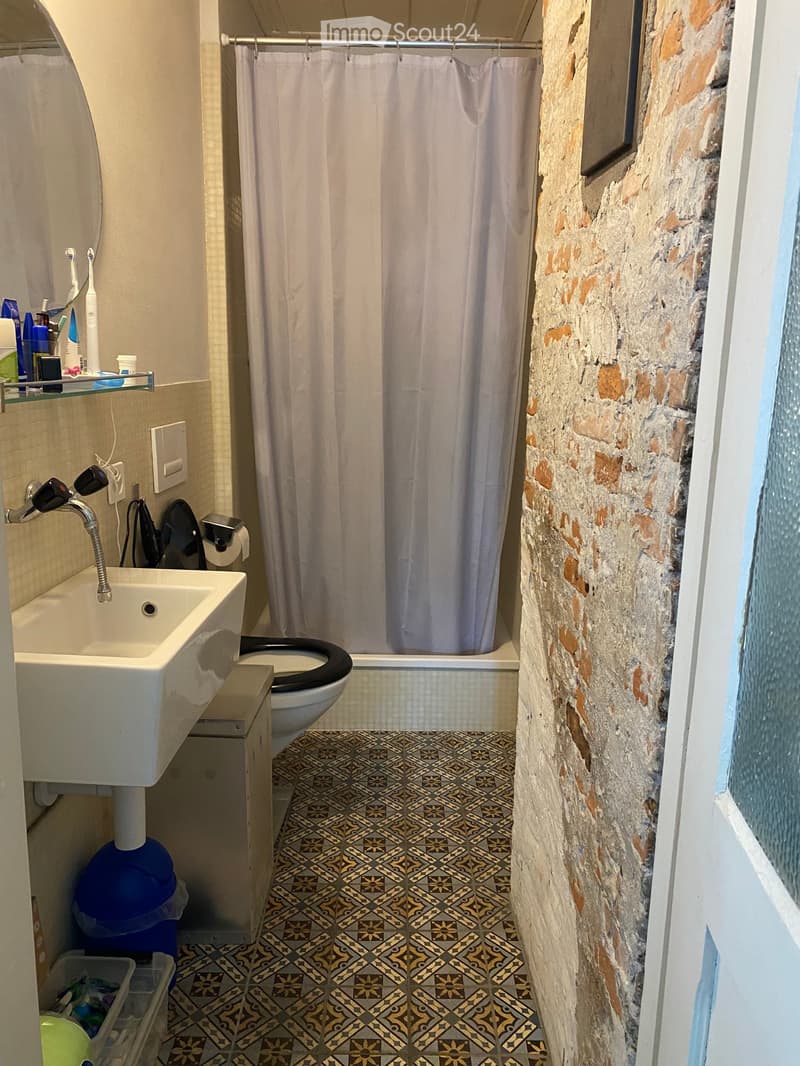 Bathroom with a sink, toilet, shower, and patterned tile floor. The shower has a curtain, and the sink has a faucet. There are toiletries on a shelf and a mirror above the sink.