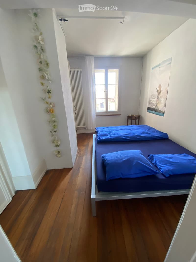 Cozy bedroom with wooden flooring, a single bed with blue bedding, a small nightstand, and a window with white curtains. A floral garland decorates the wall near the door.
