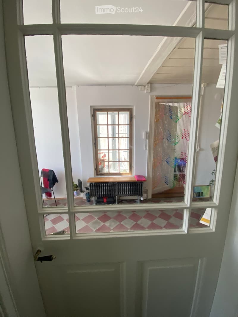 A small, furnished room with a table, radiator, chair, plant, and colorful curtain visible through an open doorway.