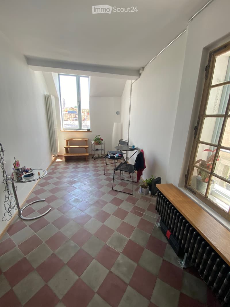 A small, well-lit room with a checkered tile floor, featuring a round table with chairs, a bench, a radiator, and a small cupboard.