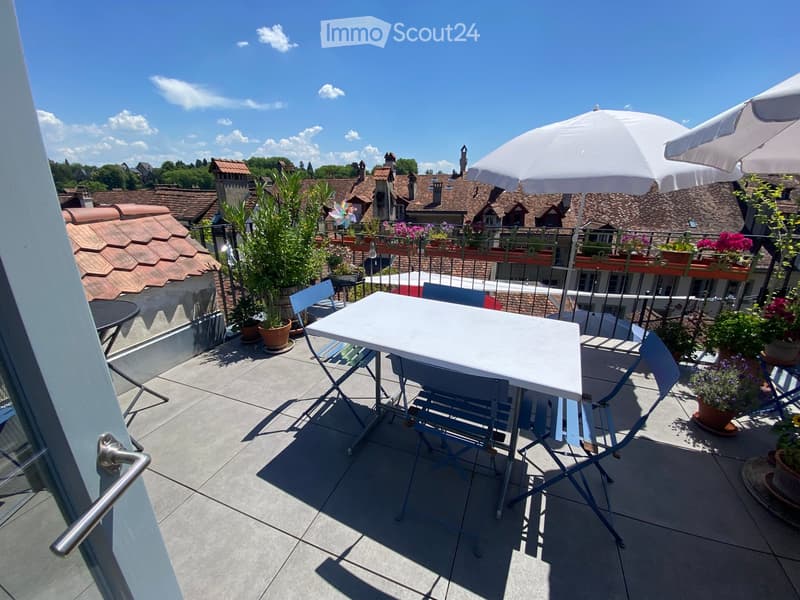 Balcony with a white table, four blue chairs, and two large white umbrellas, surrounded by potted plants and overlooking a town with red-tiled roofs.