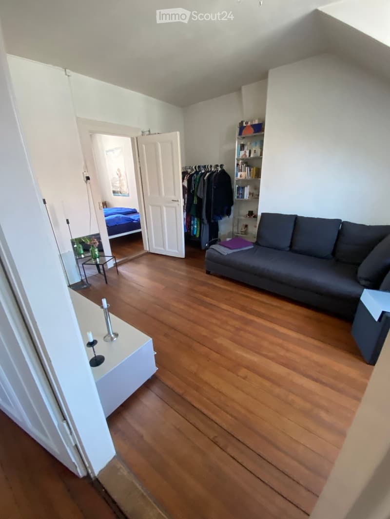 A modern living room with wooden flooring, a grey sofa, a small table with candles, a clothing rack, and an open door leading to a bedroom with a bed.