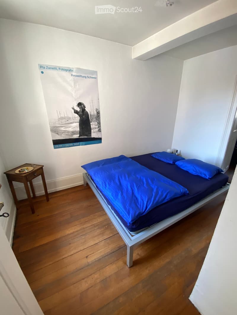 Bedroom with wooden floor, single bed with blue bedding, nightstand with a plate, and a large poster on the wall.