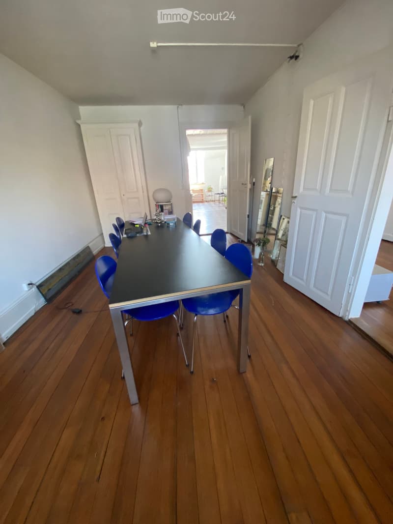 A furnished room with a long black table surrounded by blue chairs, a laptop, and a small vase with flowers. The room has wooden flooring and white walls, with a doorway leading to another room.
