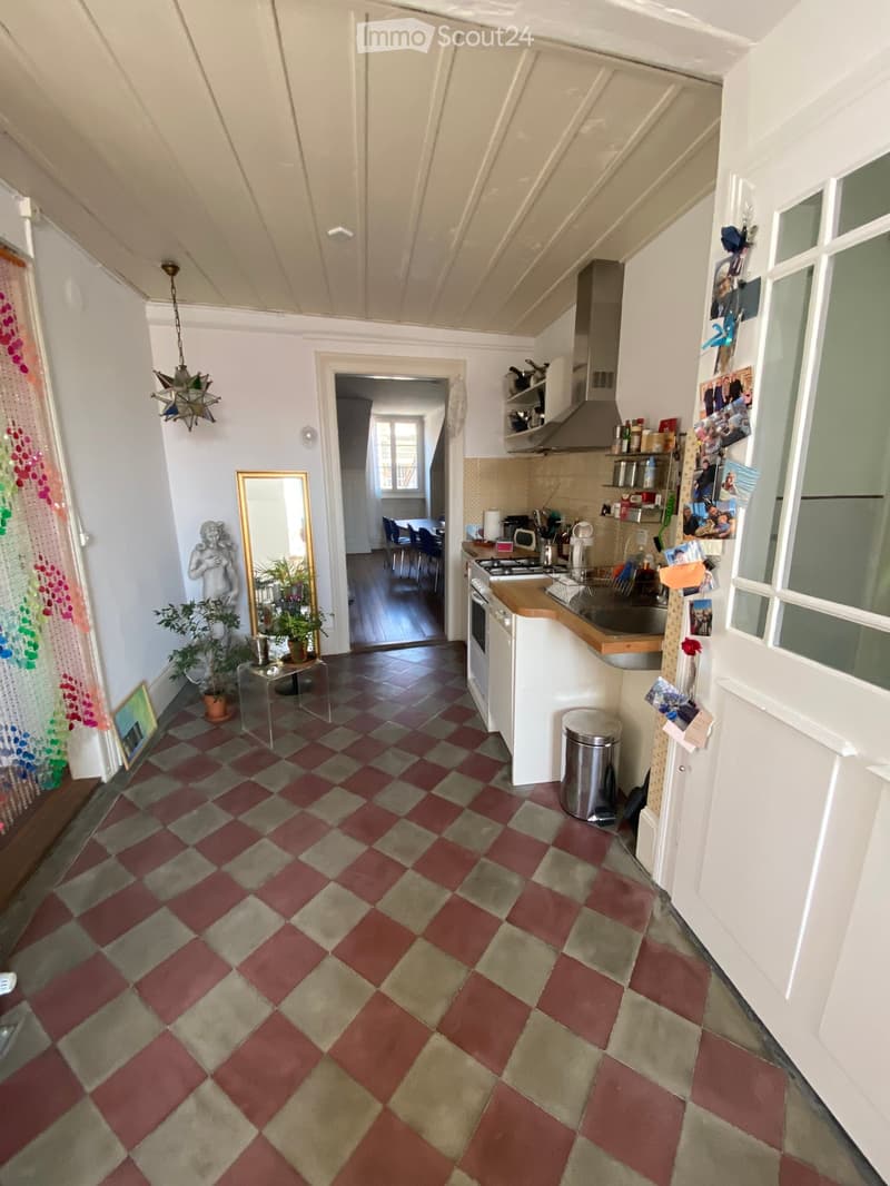 A cozy kitchen area with a checkered floor, white cabinets, and a kitchen island. Visible appliances include a stove, sink, fridge, and microwave. A small dining area with a table and chairs is visible in the background, along with a mirror, plants, and a