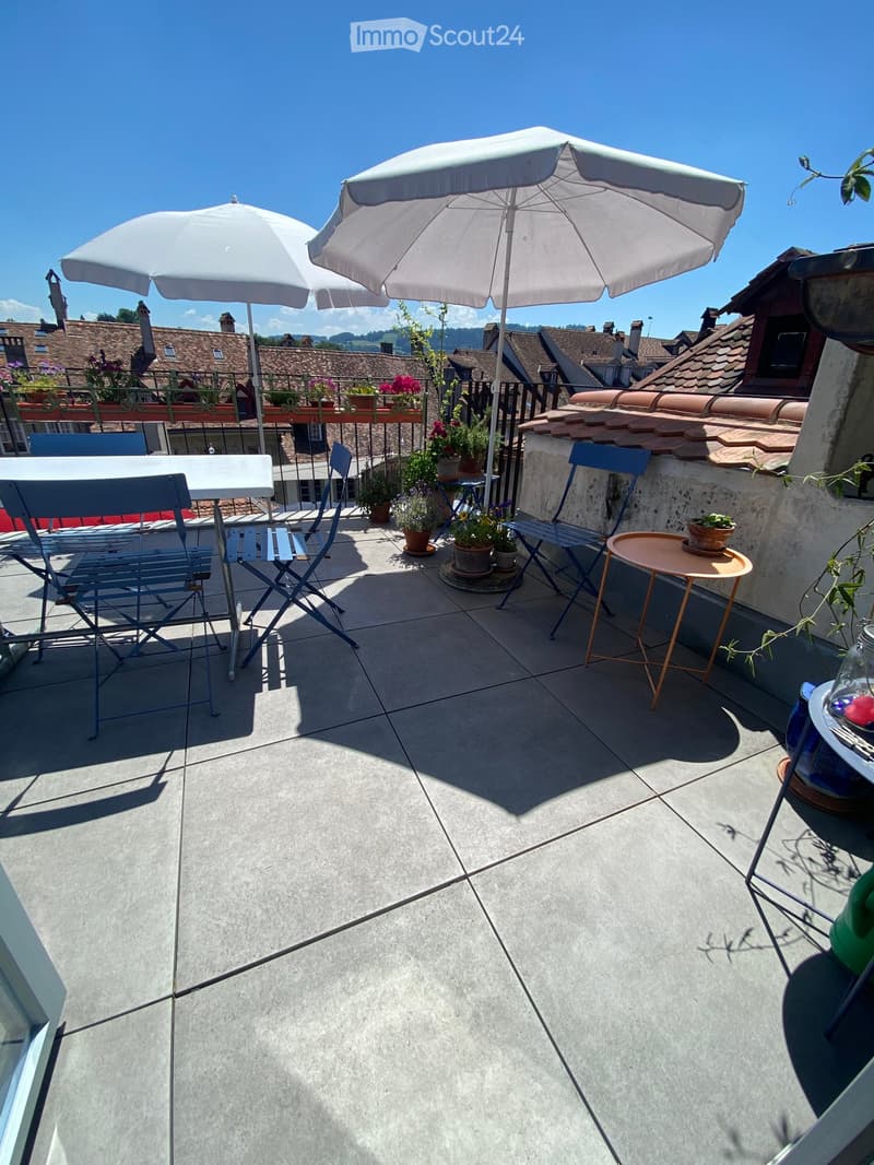 Balcony with tiled floor, two white umbrellas, a small table, chairs, and potted plants.