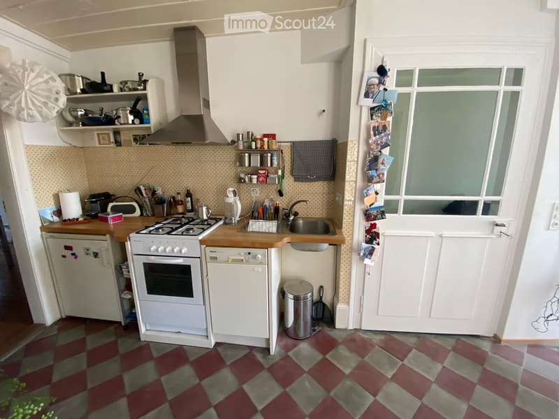 A compact kitchen with white cabinets, a gas stove, a dishwasher, and a refrigerator. The floor is tiled in a red and gray checkered pattern, and there is a window with a door leading outside.