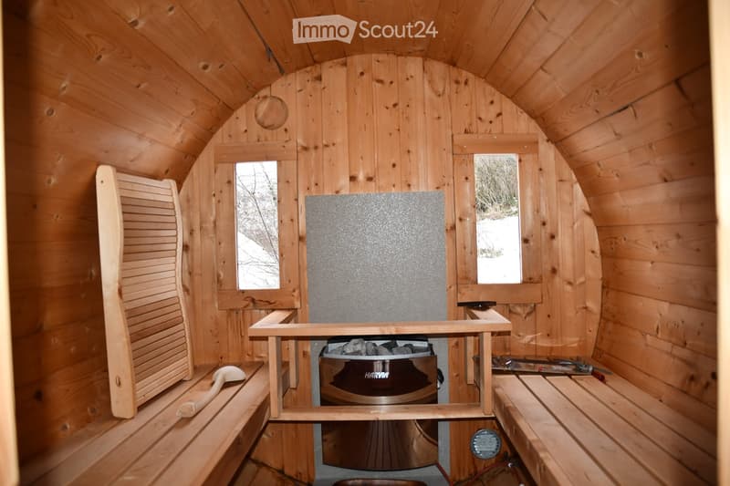 The image shows the interior of a wooden sauna with wooden benches, slats, walls, and ceiling. There are two small windows with mirrors, a wooden table, and a wooden shelf. The sauna features a central heating element with a metal surface and a small stor