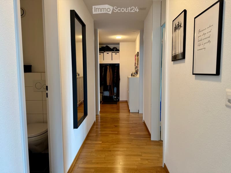 A narrow hallway with wooden flooring, white walls, and several doors leading to different rooms. There are framed pictures on the walls and a small closet with storage bins visible.