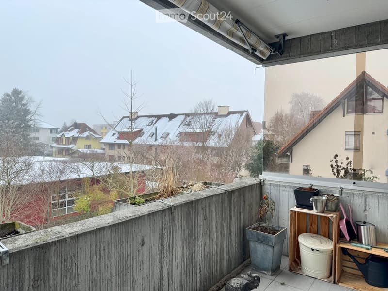 Balcony with wooden railing, potted plants, small wooden shelf holding gardening tools, and a view of snow-covered rooftops.