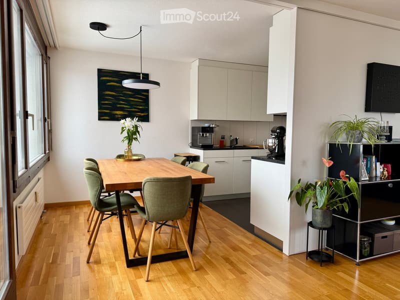 Open-plan kitchen and dining area with modern white cabinets, a wooden dining table with green chairs, and various appliances including a coffee machine and mixer. Potted plants and a painting add decor.