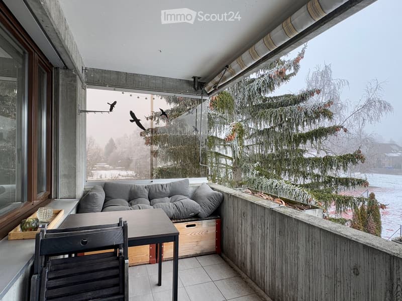 Balcony with a grey sofa, a small table, a chair, and a cup, overlooking a snowy landscape with trees.