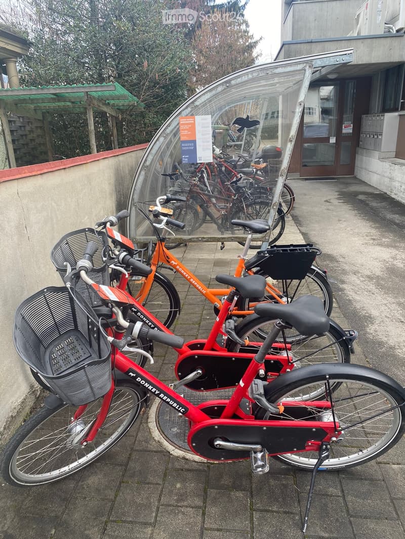 A group of red and orange electric bicycles with black baskets parked in a covered area near a building entrance.