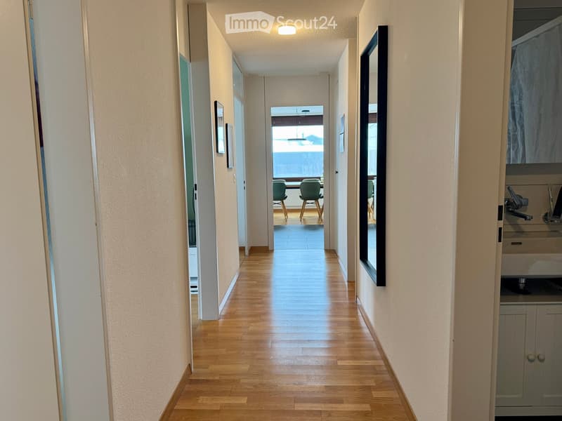 A long, well-lit hallway with wooden flooring, white walls, and a view of a room with green chairs and a table at the end.