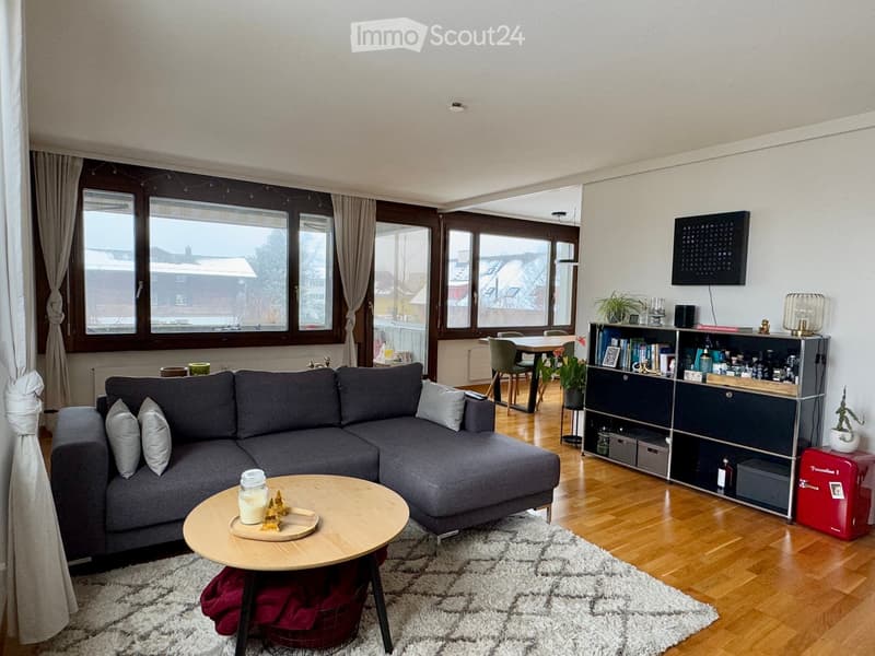 Modern living room with a grey sectional sofa, wooden coffee table, bookshelf, dining area with chairs and table, and large windows with curtains.