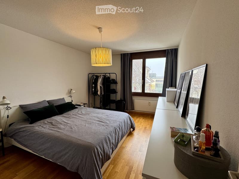 Modern bedroom with a large bed, nightstand, lamp, mirror, clothing rack, and shelf. The room features wooden flooring and large windows with curtains.