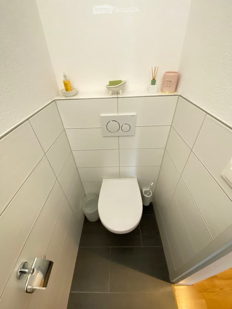 Modern, compact bathroom with white tiled walls and dark grey floor. Features a toilet, toilet paper holder, shower gel bottle, bath sponge, incense sticks, incense holder, and a cup on the shelf above the toilet.