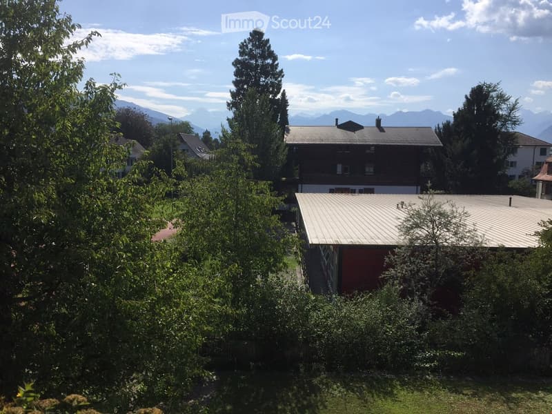 A multi-story residential building with a gray roof and white walls, surrounded by lush greenery and trees, with a scenic mountain view in the background.