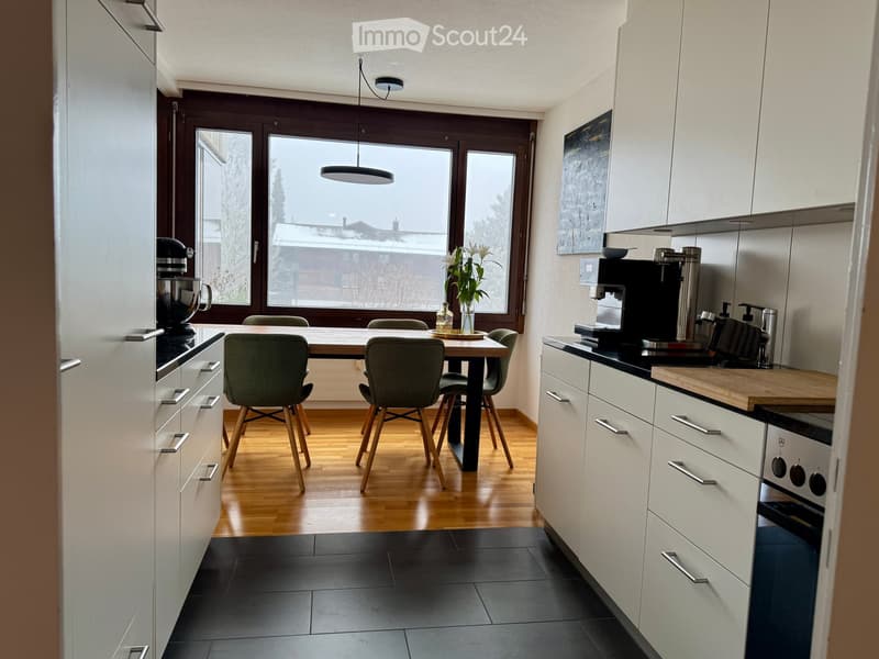 Modern kitchen with white cabinets, wooden dining table, four chairs, coffee maker, mixer, and vase with flowers on the counter.