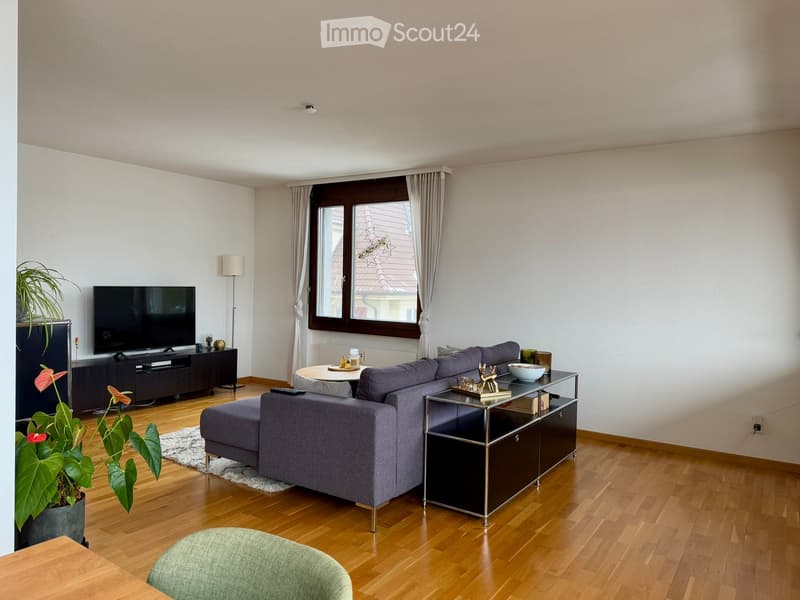 Modern living room with wooden flooring, large window with curtains, grey sectional sofa, coffee table, side table, television, floor lamp, and decorative plant.