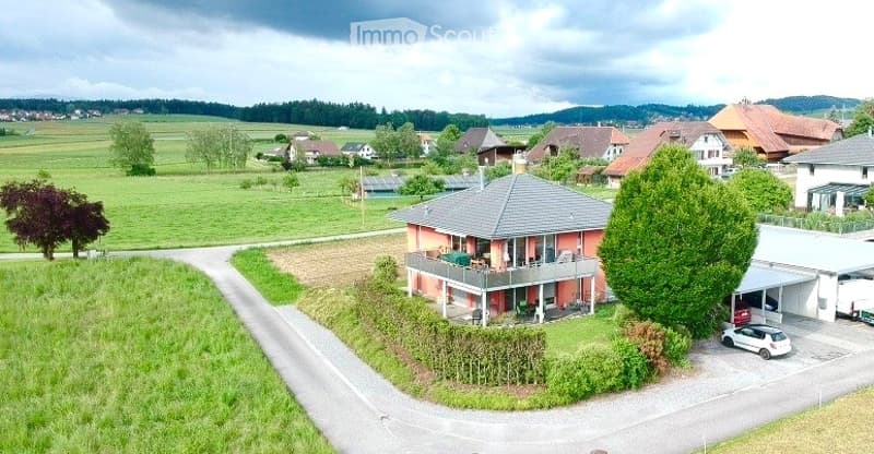 A two-story house with a modern design, featuring a garage, a parking area, and a well-maintained garden. The house is surrounded by a rural landscape with fields and trees.