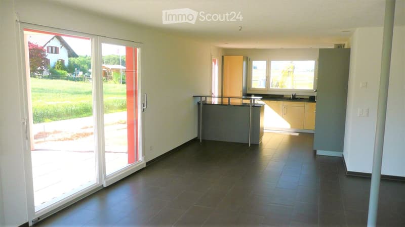 Empty kitchen with tiled floor, light-colored cabinets, and a large window. A sliding glass door leads to an outdoor area with grass and a house visible in the background.