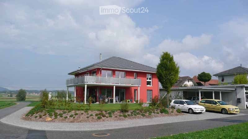 Red single-story house with a grey roof, white-framed windows, and a balcony on the front. There is a small garden area with shrubs and a driveway with two parked cars. A garage is visible to the right.
