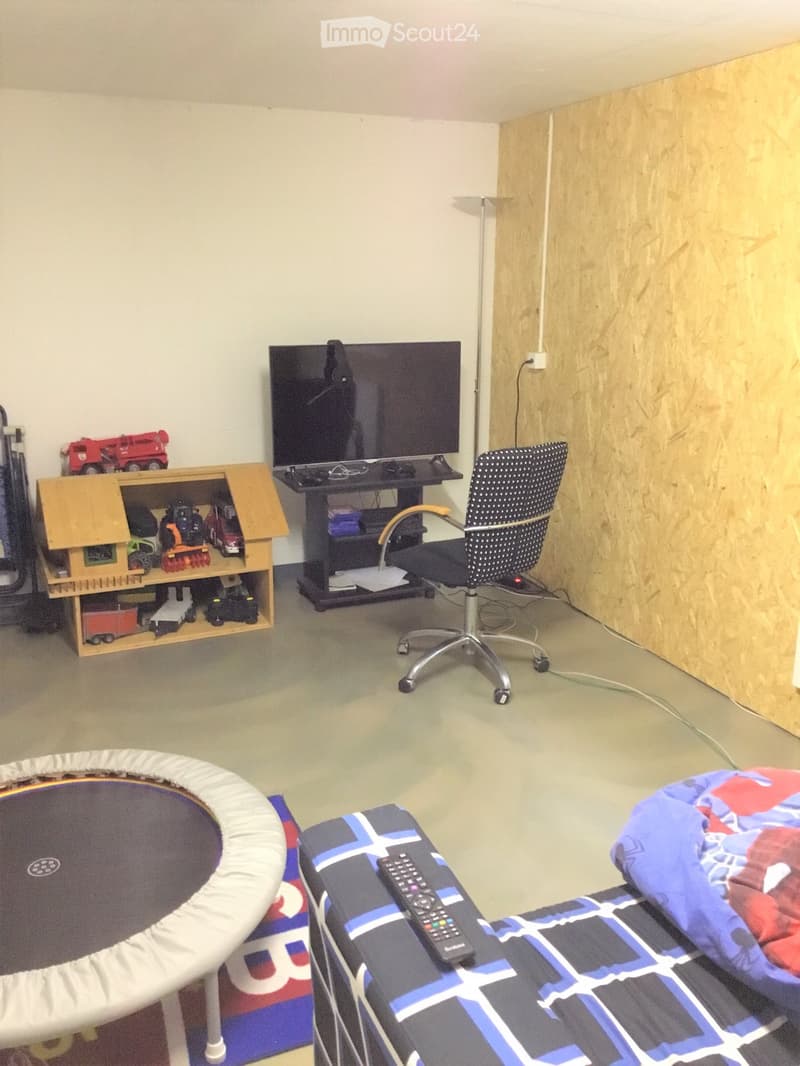 A furnished room with a chair, screen, table, shelf, remote control, toy house, toy cars, and mattress.