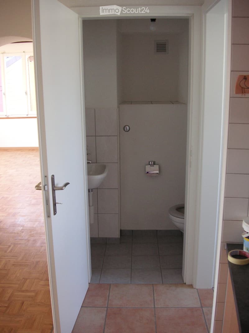A small, modern bathroom with a white sink, toilet, and tiled floor. The room has white walls and a wooden floor visible in the adjacent area.