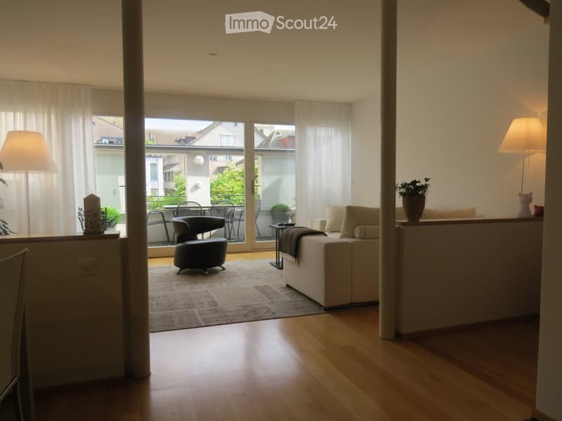 Bright living room with wooden flooring, large windows, and a balcony. Features a sofa, armchair, coffee table, and floor lamp.