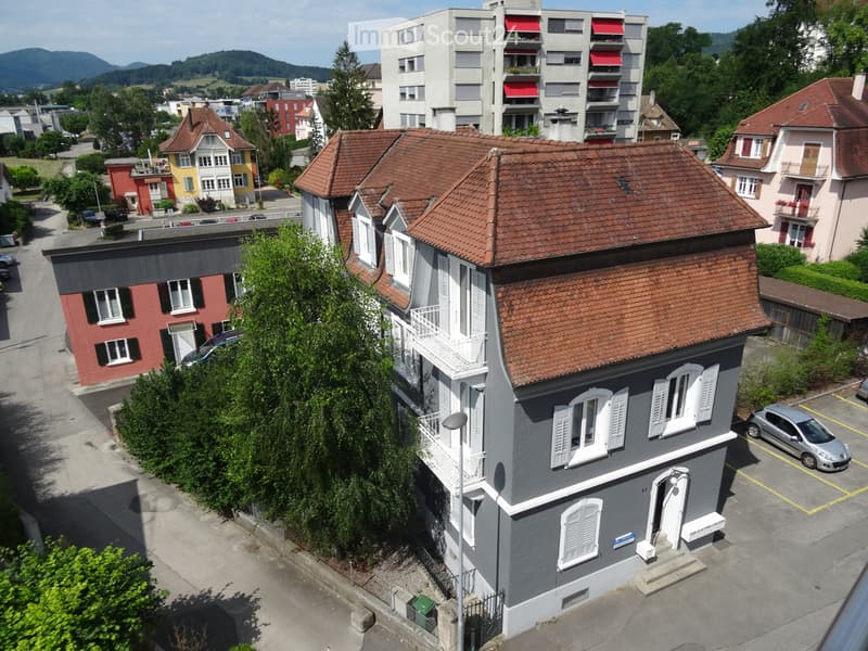 A multi-story residential building with a red-tiled roof, white-framed windows, and a grey facade. The building is situated in a neighborhood with other houses and parked cars visible in the background.