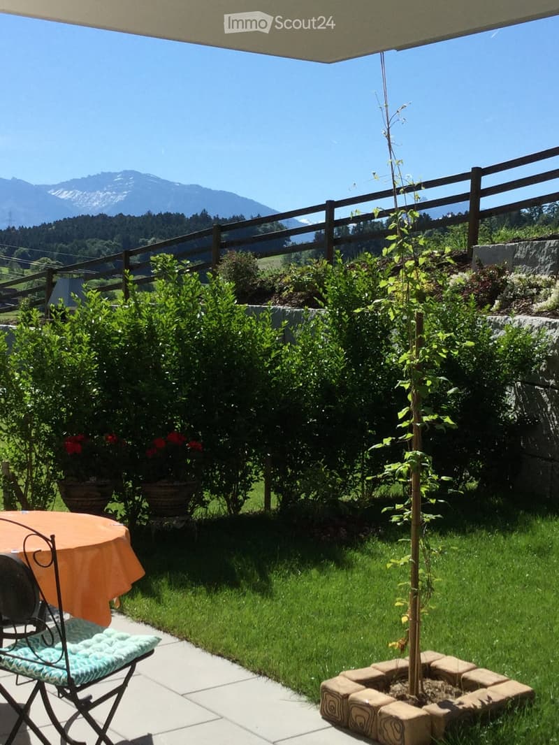 Garden view from balcony with chair, table, green bushes, small tree, stone walls, table top, red flowers.