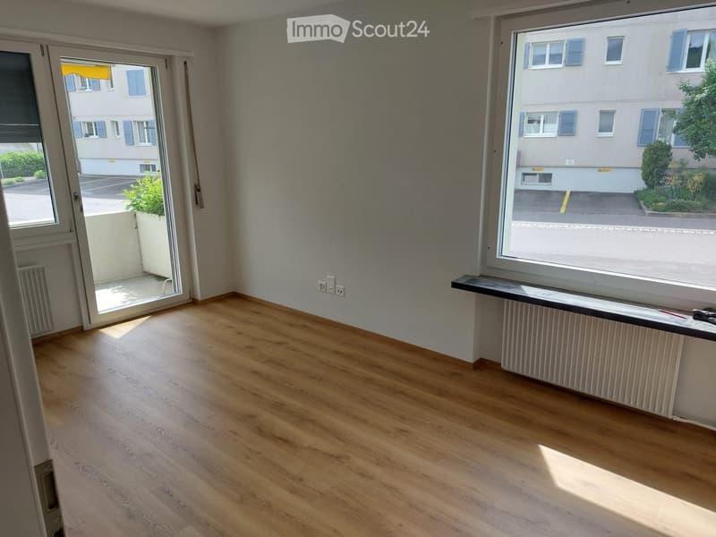 Empty room with wooden flooring, large windows, and a sliding door leading to a balcony area.
