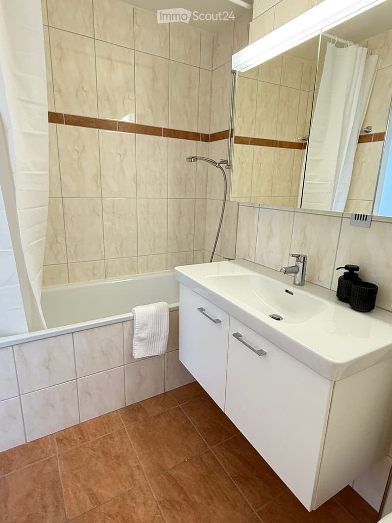 Modern bathroom with beige tiles, a bathtub, a white sink with a chrome faucet, a large mirror, and a towel on the edge of the tub.