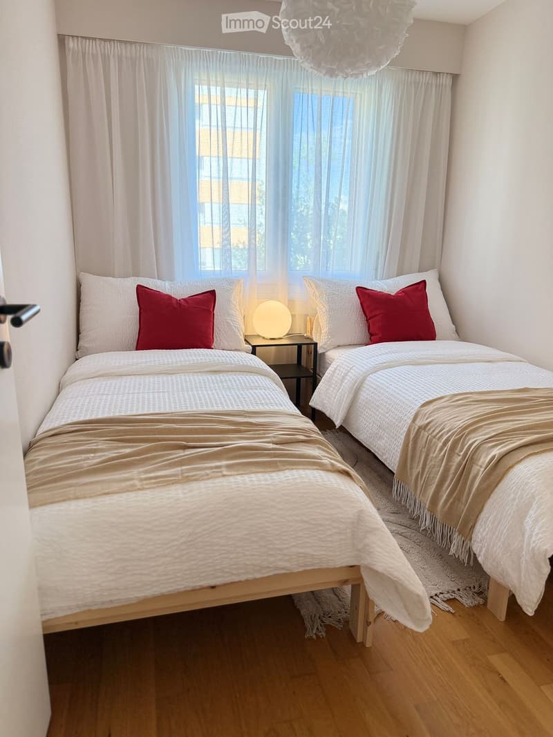 Cozy bedroom with two single beds, white bedding, red accent pillows, a small nightstand with a lamp, and large windows with sheer curtains.