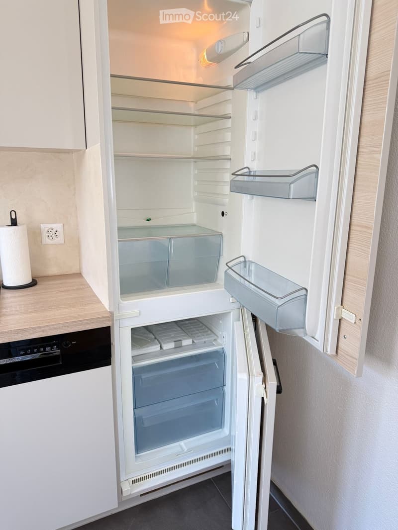 Open refrigerator with multiple compartments and shelves, adjacent to a dishwasher and a paper towel holder on the counter.