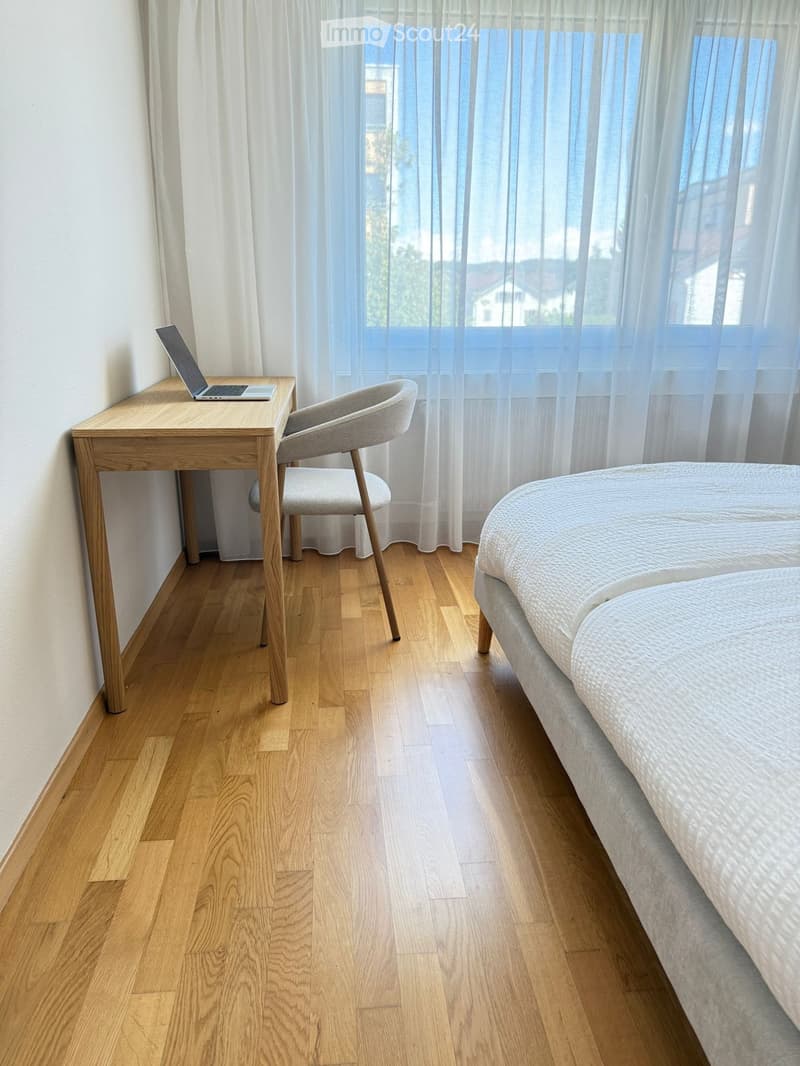 Modern bedroom with a wooden bed, a desk with a laptop, a chair, and large windows with sheer curtains.