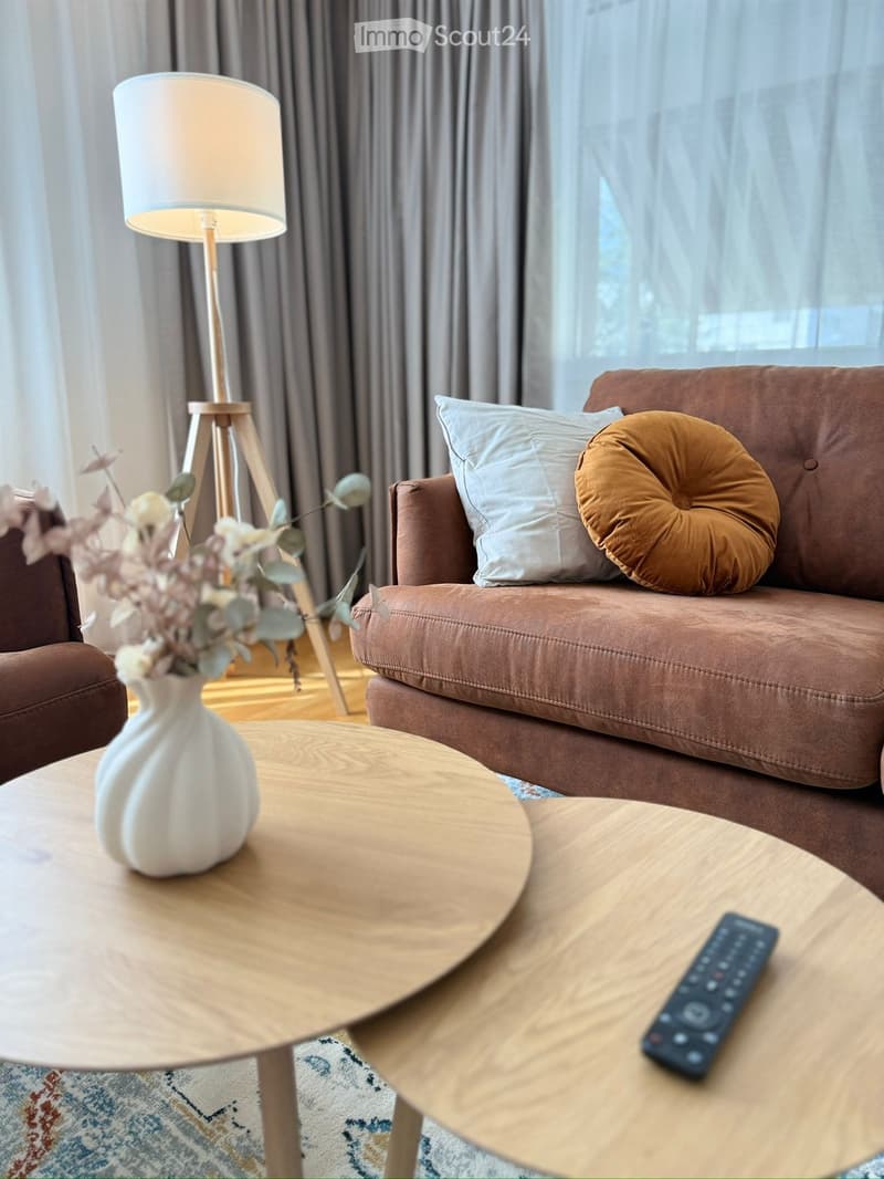 Modern living room with a brown sofa, two wooden coffee tables, a white vase with flowers, and a floor lamp.