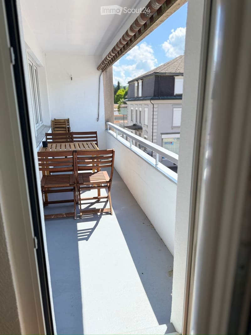 Balcony with wooden table and chairs, white walls, metal railing, and view of neighboring buildings.