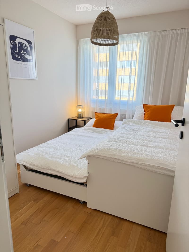 Modern bedroom with a white bed, orange pillows, nightstand, table lamp, and a large window with sheer curtains.