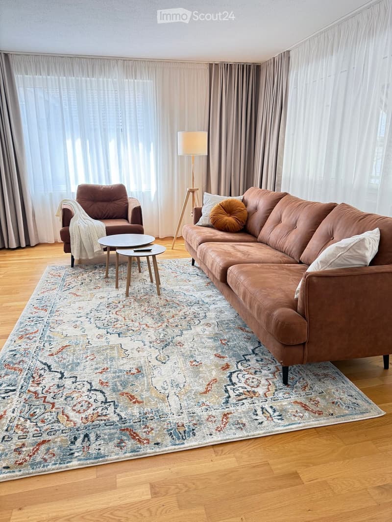 Modern living room with wooden flooring, large windows with sheer curtains, a brown leather sofa, armchair, coffee table, floor lamp, and decorative throw blanket and pillows.