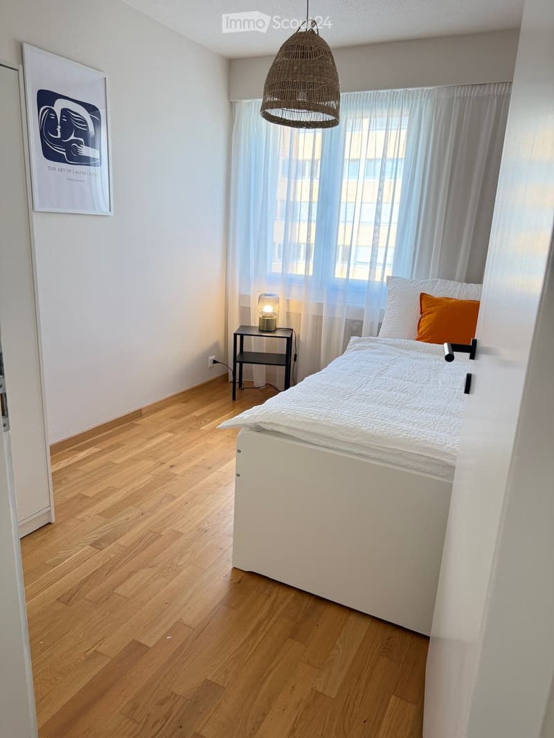 Cozy bedroom with wooden flooring, a white bed with white and orange pillows, a nightstand with a lamp, and a large window with sheer curtains. A framed picture hangs on the wall.