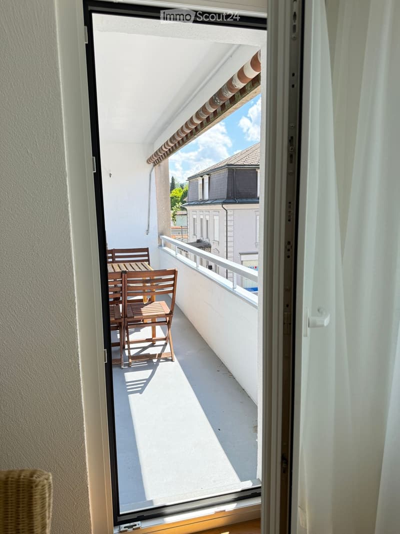 Balcony with a table and chairs, overlooking a building with a clear sky and some greenery.